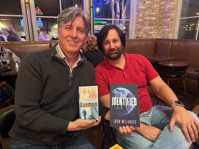 Daniel Suarez and John sitting in a bar. Daniel holds up his novel 