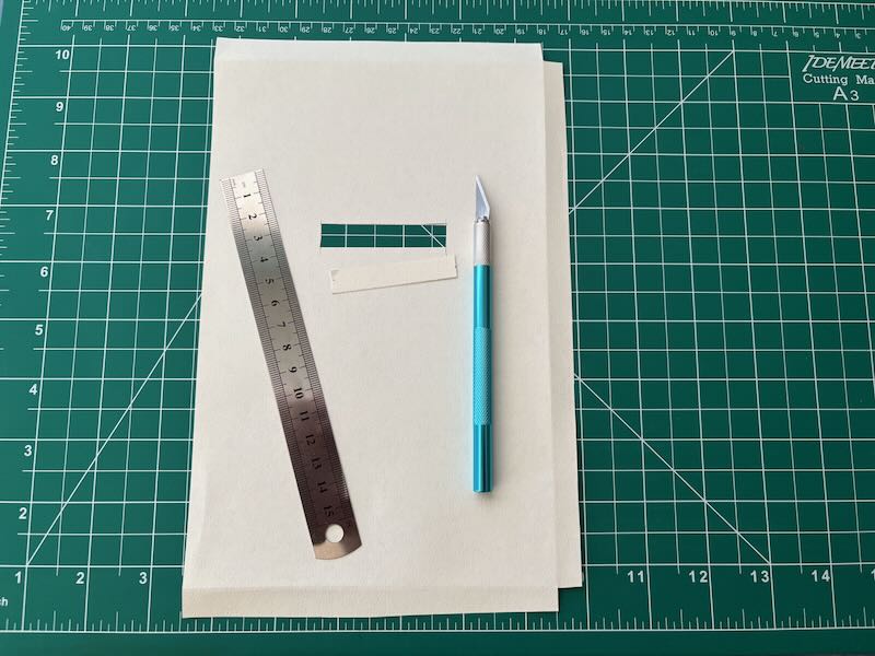An X-Acto knife, a metal ruler, and a thick piece of paper with a cut-out horizontal window where the binary numbers will be stamped.