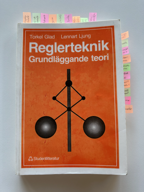 Photo of the book "Reglerteknik: Grundläggande teori" which is from a Swedish university class in automatic control. It has about 20 sticky index tabs in pastel colors sticking out from the top and side.