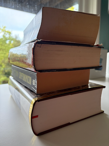 John's stack of four thick books that trouble him, laying on a white desk.